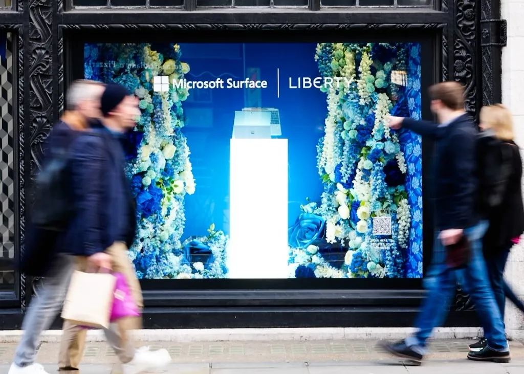 A shop window display showcasing the Microsoft Surface and Liberty collaboration. The background features decorative blue and white flowers. Two people walk past the window, one holding a shopping bag.