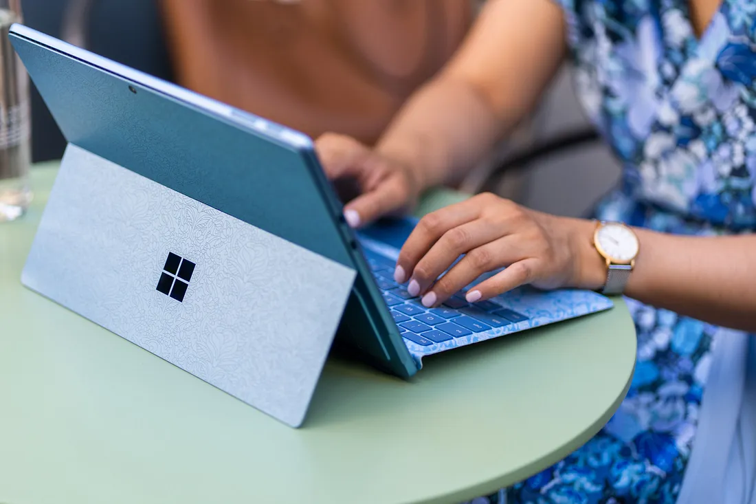 A person wearing a floral dress types on a blue tablet with a detachable keyboard on a green table. The tablet has a textured design on the back and a small, black square logo. The person is also wearing a gold watch.