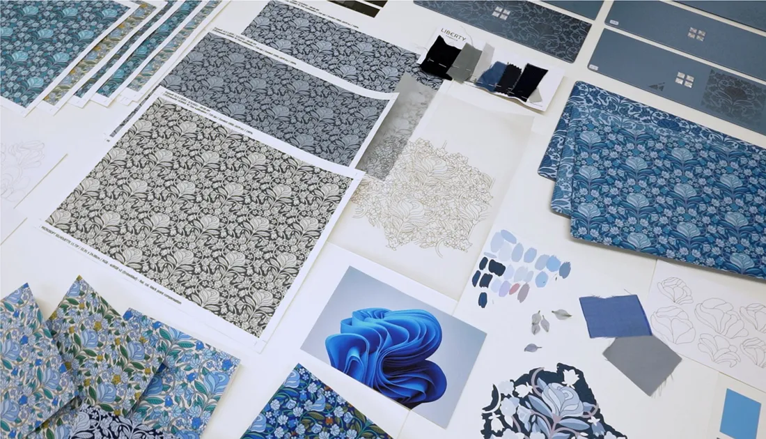 A collection of floral pattern samples and color swatches arranged on a table. The designs feature intricate blue and gray floral motifs, along with paint chips and digital illustrations, showcasing a creative design process.