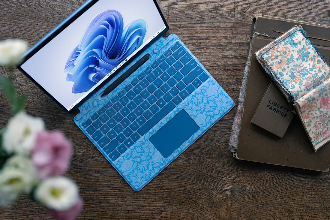 A blue laptop with a floral pattern on the keyboard rests on a wooden table. The screen displays a modern operating system. Nearby are floral-patterned notebooks and a stack of old books. A vase with pink and white flowers is in the foreground.