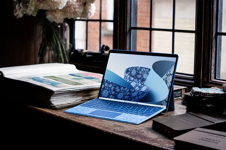 A sleek laptop with a decorative blue design sits on a wooden desk near a large window. The desk is adorned with an open book, a vase of flowers, and several closed notebooks, creating a cozy, sunlit workspace ambiance.