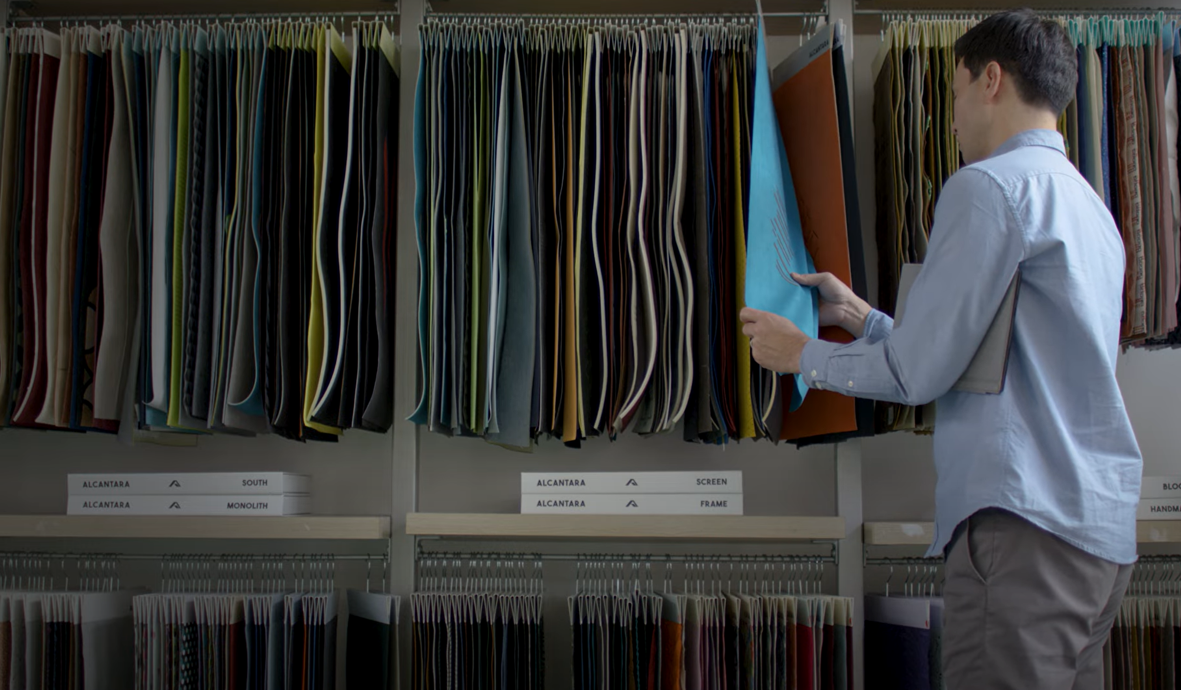 A person stands in front of shelves filled with hanging fabric samples, flipping through them. The shelves have labels, and the fabrics are in various colors and textures. The person wears a light blue shirt and beige pants.