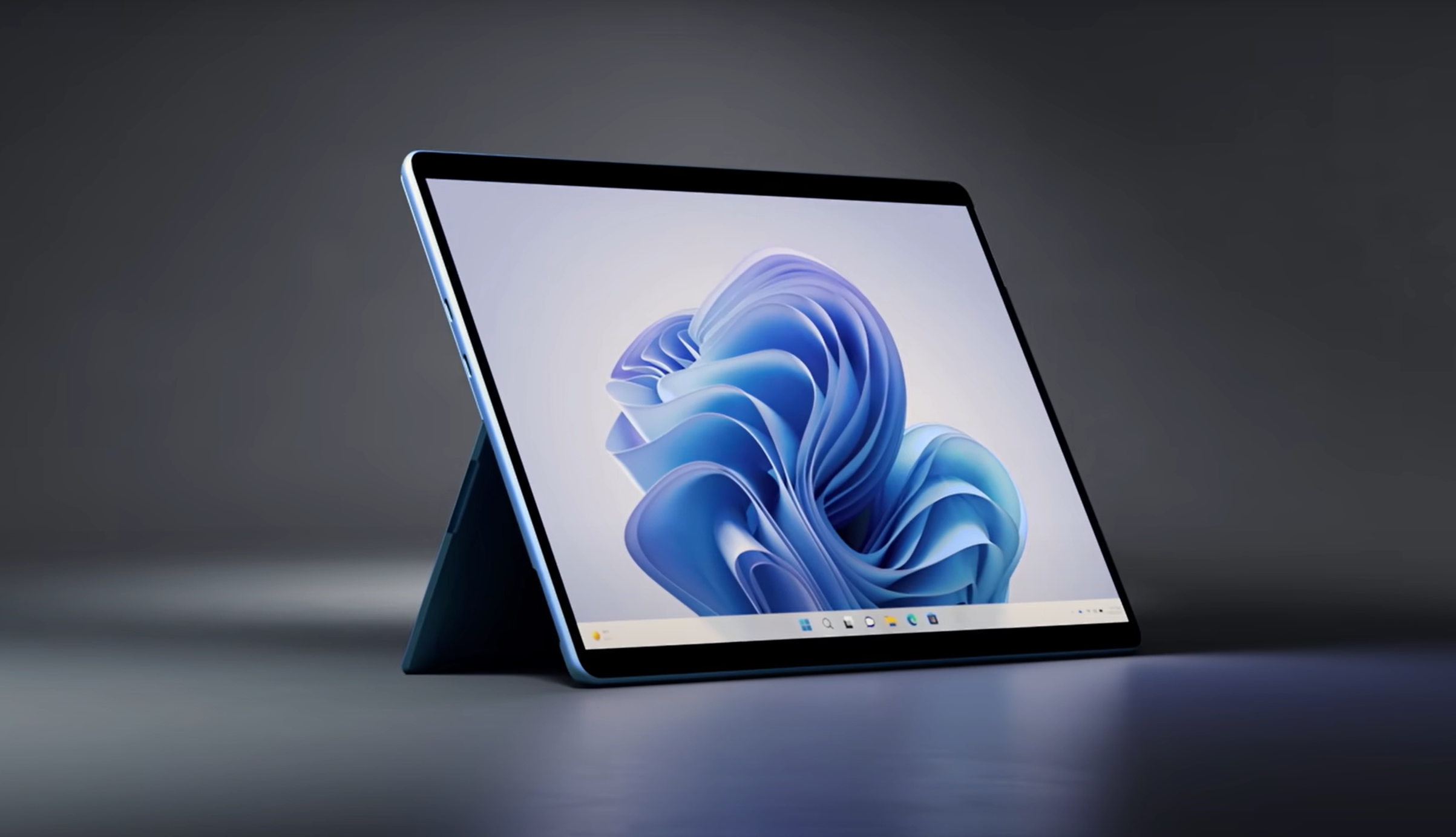 A sleek tablet with a detachable keyboard stands on a surface, displaying a blue abstract design on its screen. The background is a dark, blurred gradient, highlighting the device's modern and minimalist design.