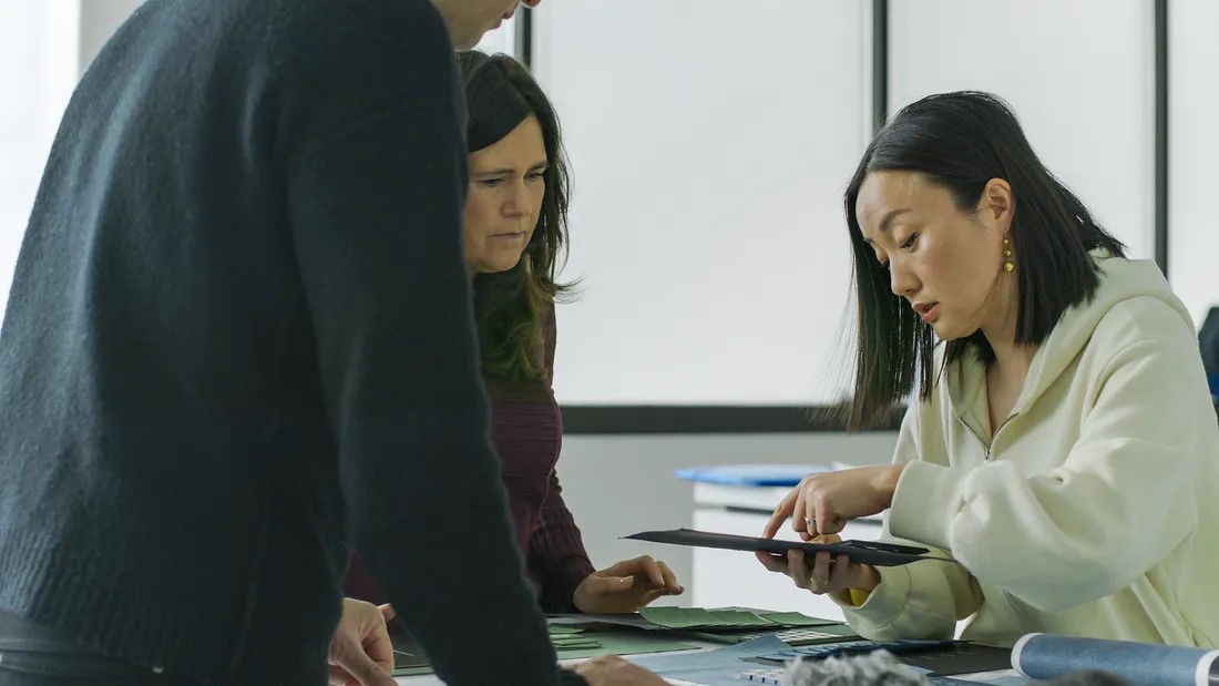 Three people are collaborating at a table, examining documents and materials. One person standing on the left has their back turned, while the two others, a man and woman, look at a folder the woman in white is showing. Professional setting with natural light.