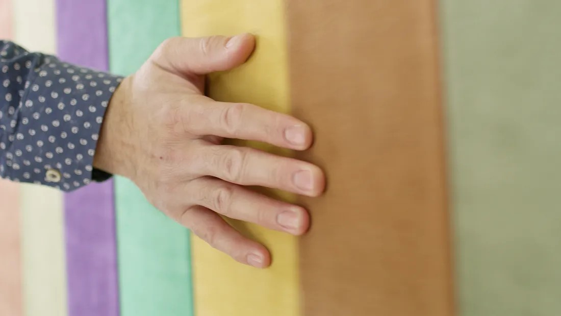 A hand with a blue polka-dotted shirt sleeve is shown resting on a colorful wall with vertical stripes in green, yellow, orange, brown, purple, and beige.