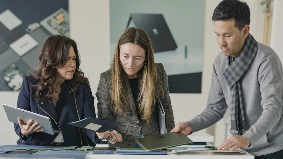 Three people are gathered around a table, looking at various fabric and material samples. The person on the left is holding a tablet and fabric sample, the person in the middle is examining a fabric, and the person on the right is reviewing material samples on the table.