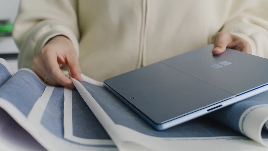 A person wearing a light-colored jacket is holding a blue laptop with one hand and flipping through a large blueprint with the other. The laptop has a reflective Windows logo on the back. The scene appears to be in an office or workspace.