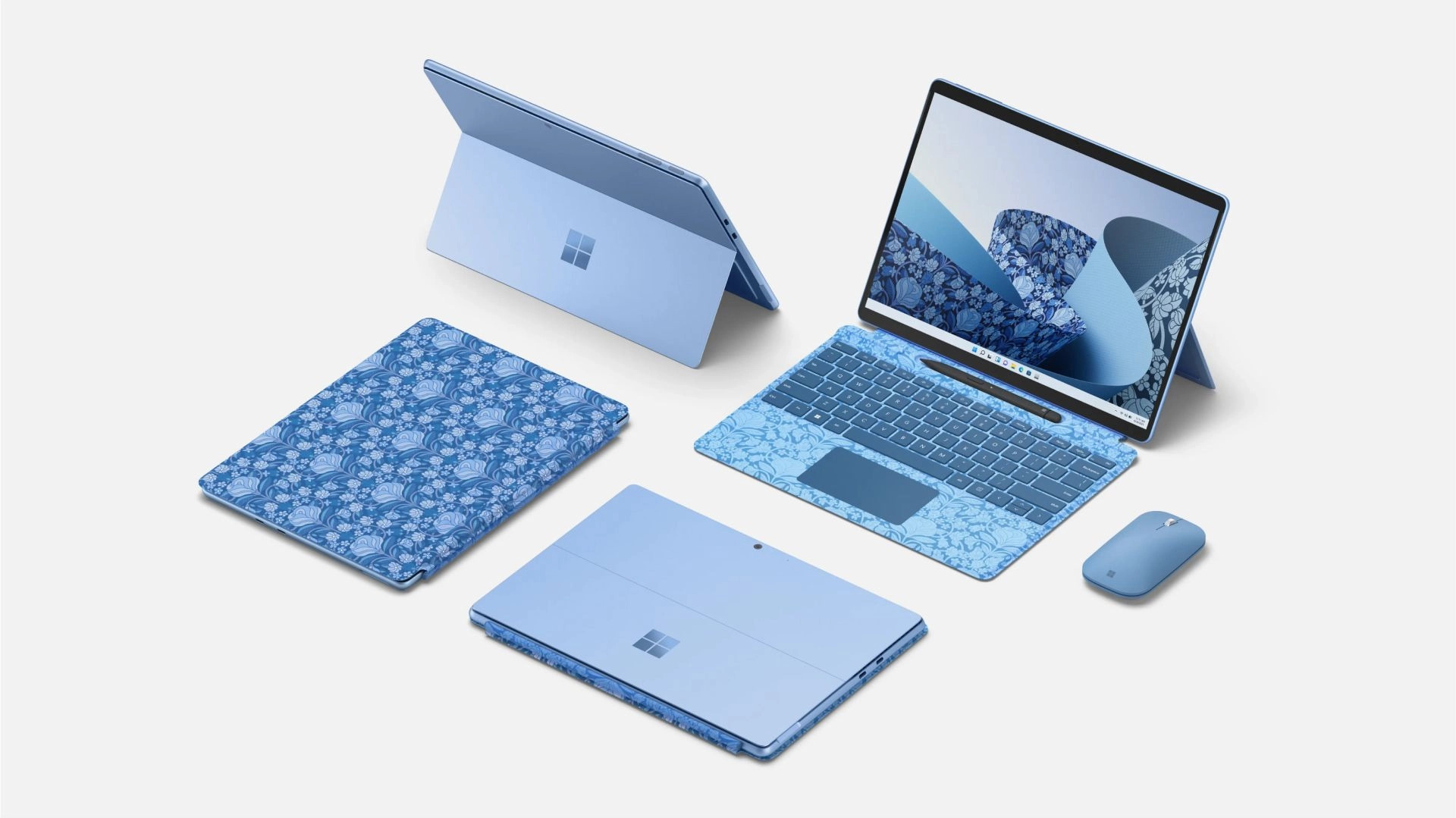 A group of Microsoft Surface devices in various shades of blue are displayed on a white background. The set includes a laptop, a tablet, a Bluetooth mouse, and two tablets with patterned covers featuring a floral design. The laptop screen shows a blue abstract pattern.