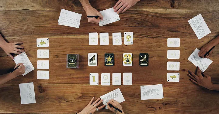 People sitting around a wooden table playing a card game. Various cards with illustrations and numbers are spread across the table, surrounded by handwritten notes. Hands holding pens and writing on note cards are visible.