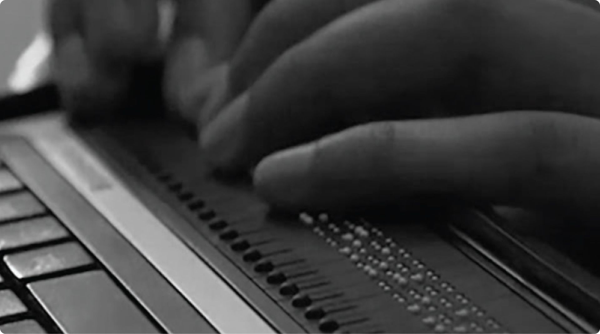 A close-up, black-and-white photo of hands using a braille display keyboard, which has raised dots for tactile reading input, connected to a laptop.