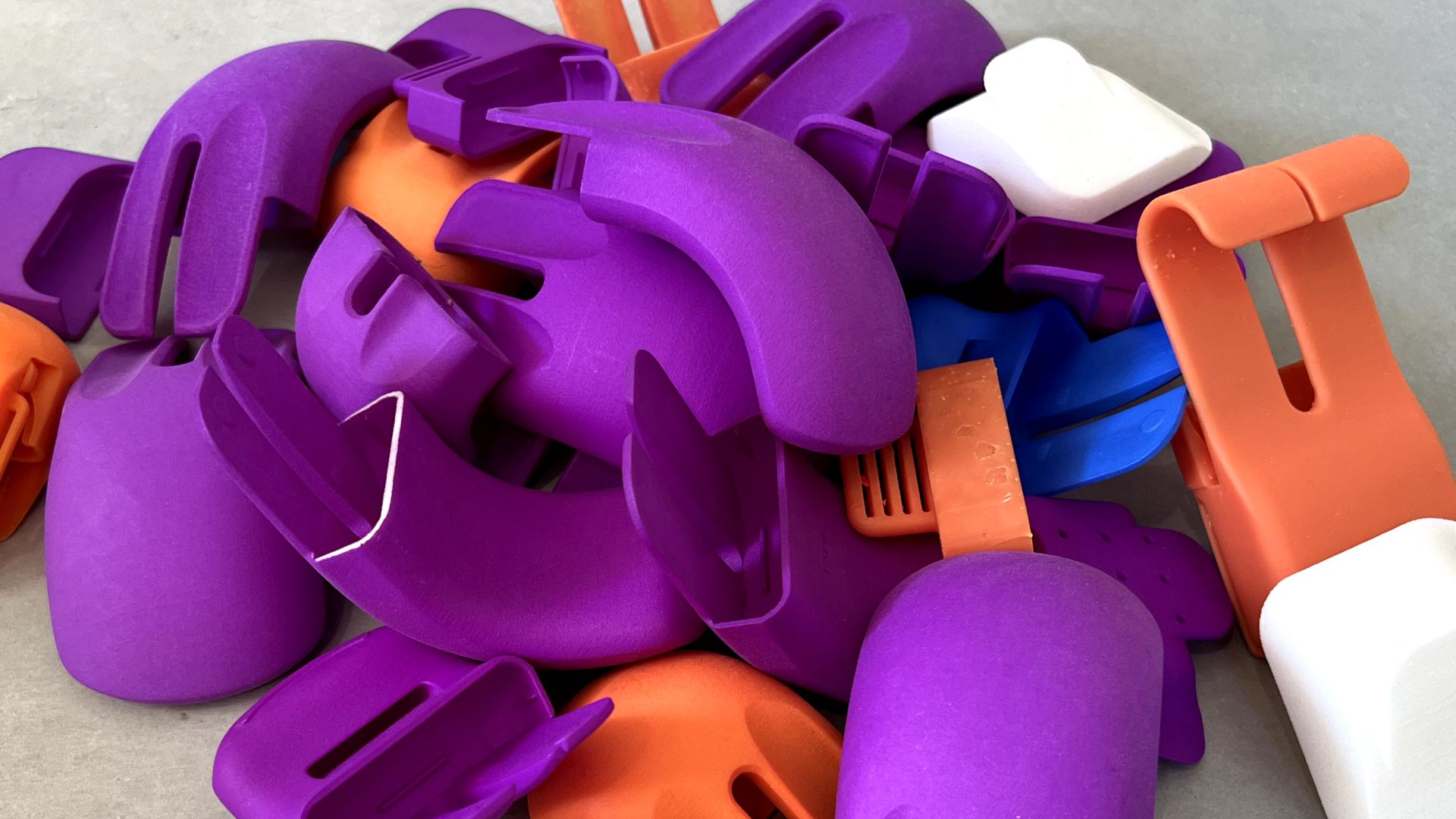 A pile of brightly colored plastic rock climbing holds, mainly in purple, orange, and white, scattered on a gray surface. The holds vary in shape and size, designed for different types of climbing grips.