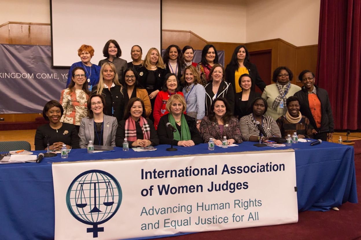 A group of women, including international judges, are gathered for a group photo in front of a banner that reads "International Association of Women Judges: Advancing Human Rights and Equal Justice for All." Some are seated at a table, while others stand behind them.