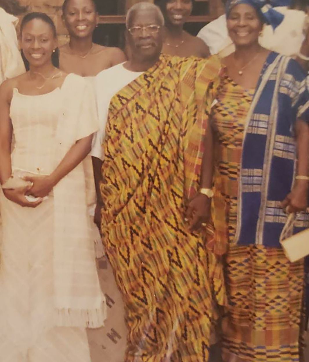 A group of people are gathered, with three people prominently in the front center. The person in the middle is wearing a traditional kente cloth, flanked by a woman in a white dress on the left and a woman in a blue and yellow patterned outfit on the right.