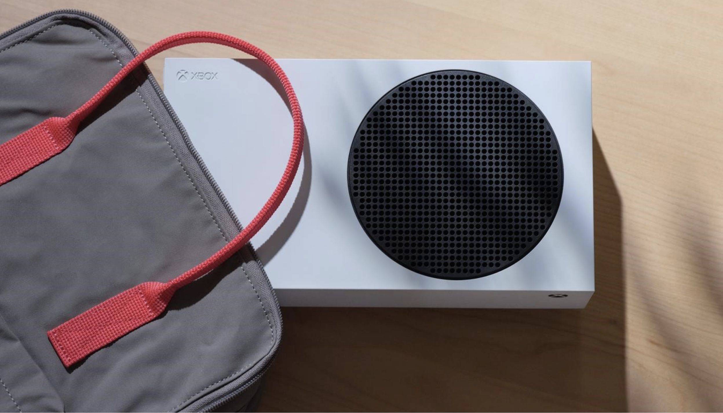 A white Xbox console with a black circular vent is placed on a light wooden surface. Next to the Xbox is a gray bag with red straps.