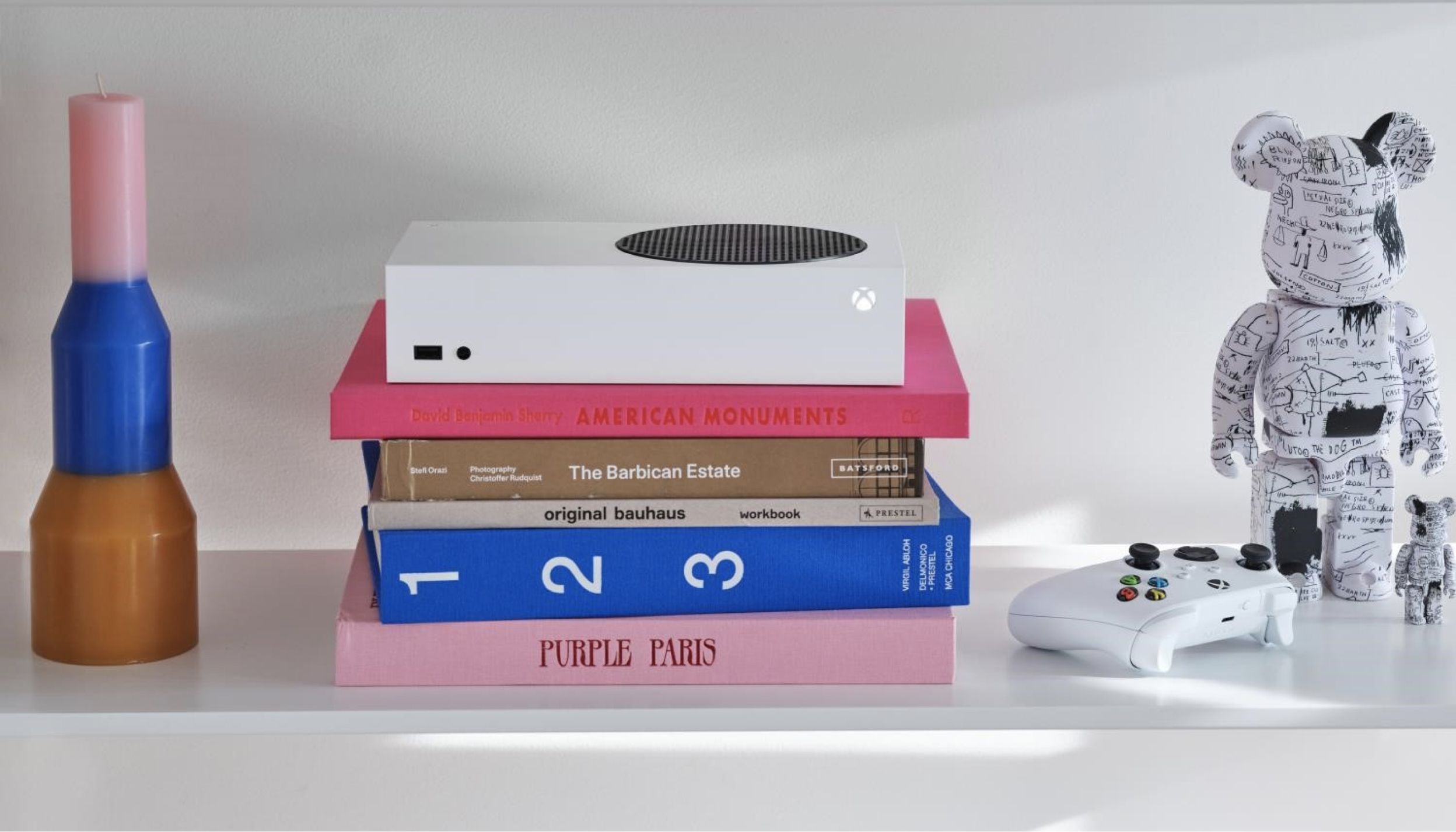 A neatly organized shelf displays a white game console, a white game controller, a multicolored vase, a stack of books with various titles, and a decorative bear figurine with a black and white pattern.