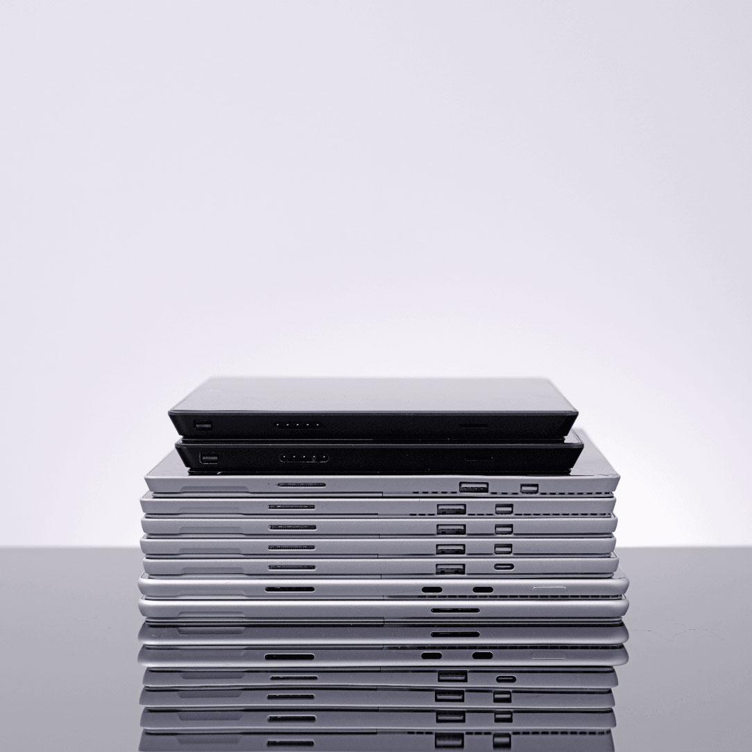 A neat stack of various laptops arranged on top of one another against a plain background. The laptops are in different colors, primarily silver, gray, and black, highlighting their thin, sleek designs.