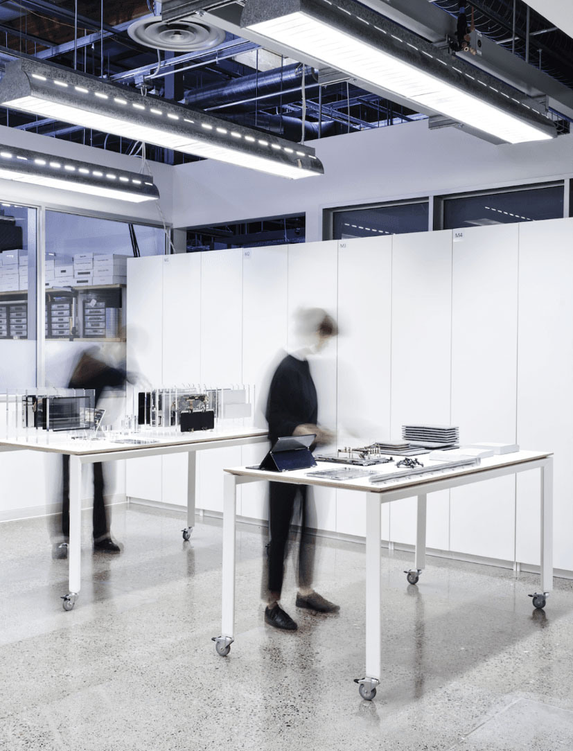 A modern, well-lit office space with blurred figures of people moving around two tables displaying various documents and small models. The room has a polished concrete floor, large cabinets, and ceiling lights, creating a clean and organized atmosphere.