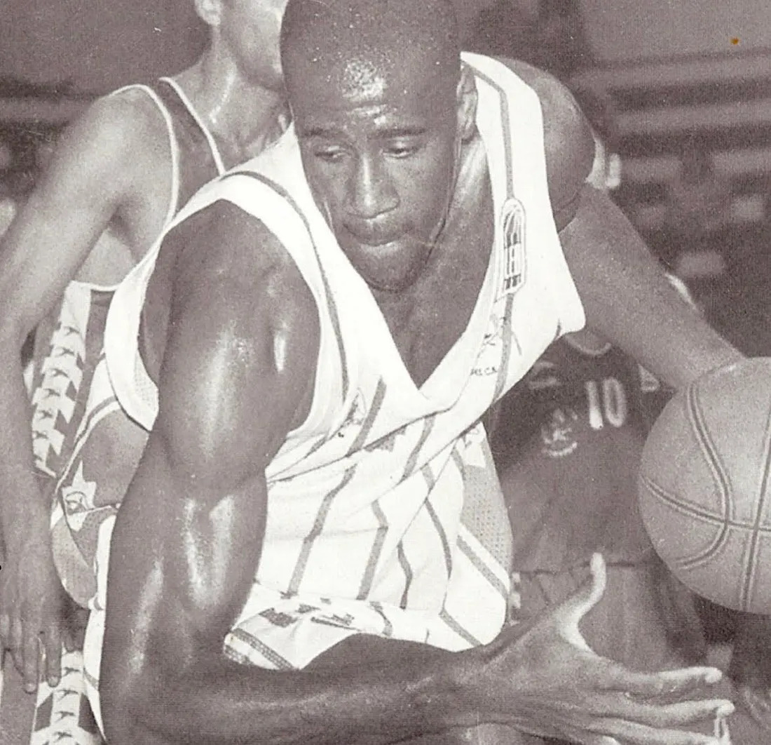 Black and white photo of a basketball player in a sleeveless uniform, dribbling a basketball with one hand while looking downward. Another player in a contrasting uniform is seen in the background, partially obscured.