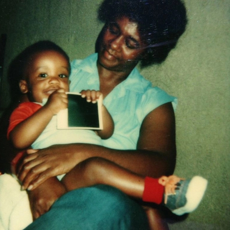 A woman lovingly holds a young child on her lap. The child, wearing a red and white shirt and red socks with blue shoes, is smiling and holding up a small rectangular object. The woman, in a light blue sleeveless blouse, looks down at the child with a gentle expression.