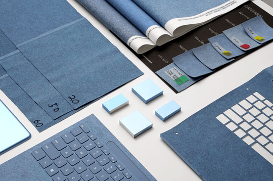 A flat lay image of various office supplies in shades of blue and white, including a laptop with a blue keyboard, sticky notes, paper samples, and fabric swatches. The items are neatly arranged on a white surface, creating a visually appealing and organized layout.