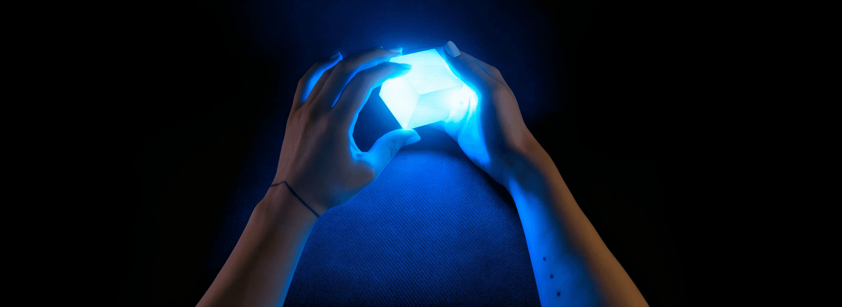 Two hands gently hold a glowing blue cube against a dark background. The light from the cube illuminates the hands and creates a soft blue glow around them. The image has a mysterious and futuristic feel.