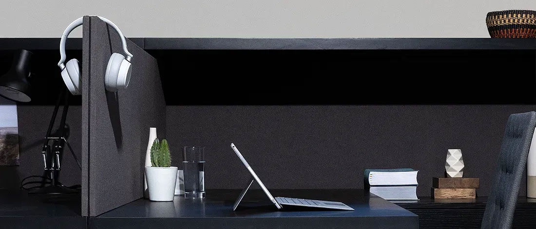 A modern workspace features a white tablet set up on a dark desk, flanked by a potted cactus, a glass of water, and a desk lamp with white headphones hanging on a divider. In the background, there are shelves with stacked books and a decorative vase.