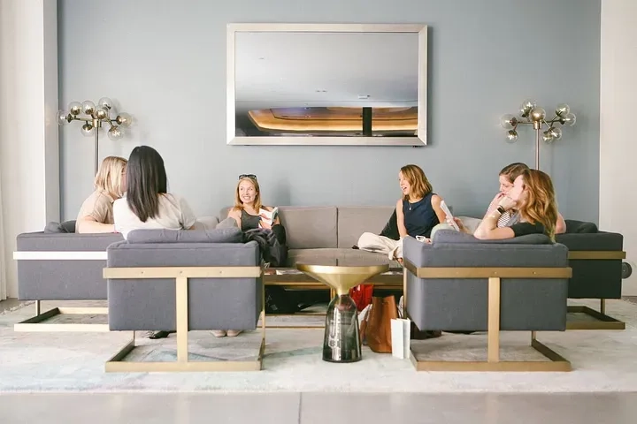 A group of six individuals are sitting on modern gray couches arranged in a square in a contemporary living room. Behind them, a large mirror hangs on the wall. They appear to be engaged in conversation, some holding drinks, with a golden coffee table in the center.