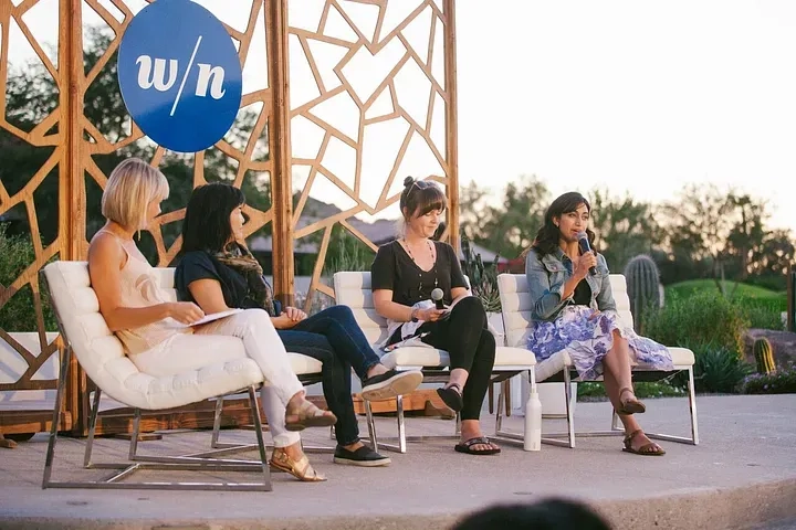 A group of four women sit on a stage having a panel discussion outdoors. The woman on the far right is holding a microphone while the others listen attentively. They are seated in white chairs with a geometric wooden backdrop behind them and trees in the background.