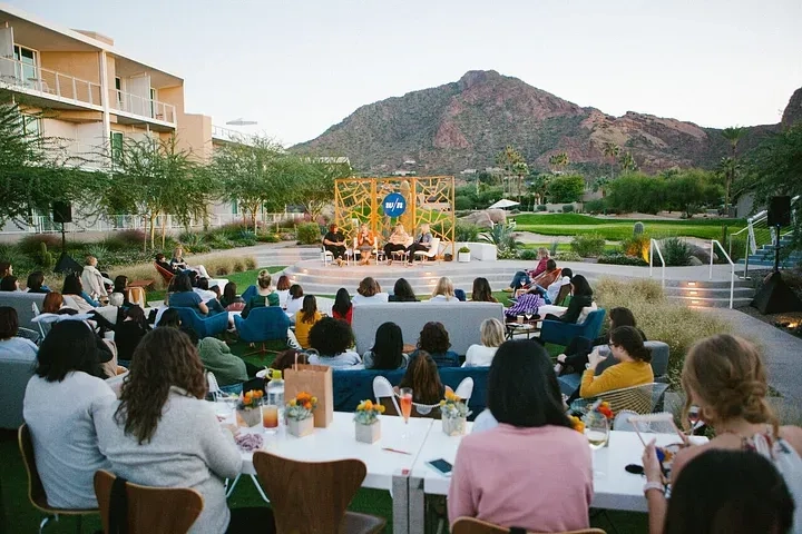 Outdoor conference with attendees seated in a semi-circle facing a stage with a panel of speakers. The backdrop includes a lush garden and mountainous landscape in the distance. A modern building and green lawn are visible.