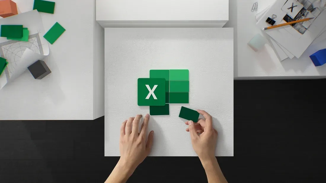 A pair of hands arranges green square cards on a white surface, with the top card displaying a white "X" symbol, resembling the Microsoft Excel logo. Nearby are office supplies, including grid paper, pens, and small objects scattered around the desk.