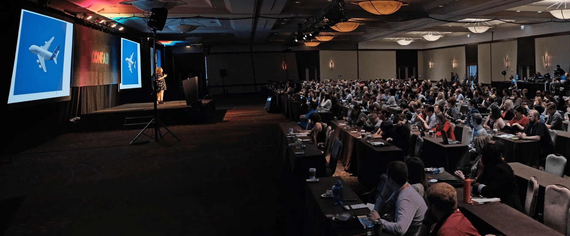 A speaker stands on a stage presenting during a conference. A large screen displays an image of an airplane in flight. The audience, seated in rows, attentively listens and watches. The room is dimly lit, with overhead lights and spotlights illuminating the stage.