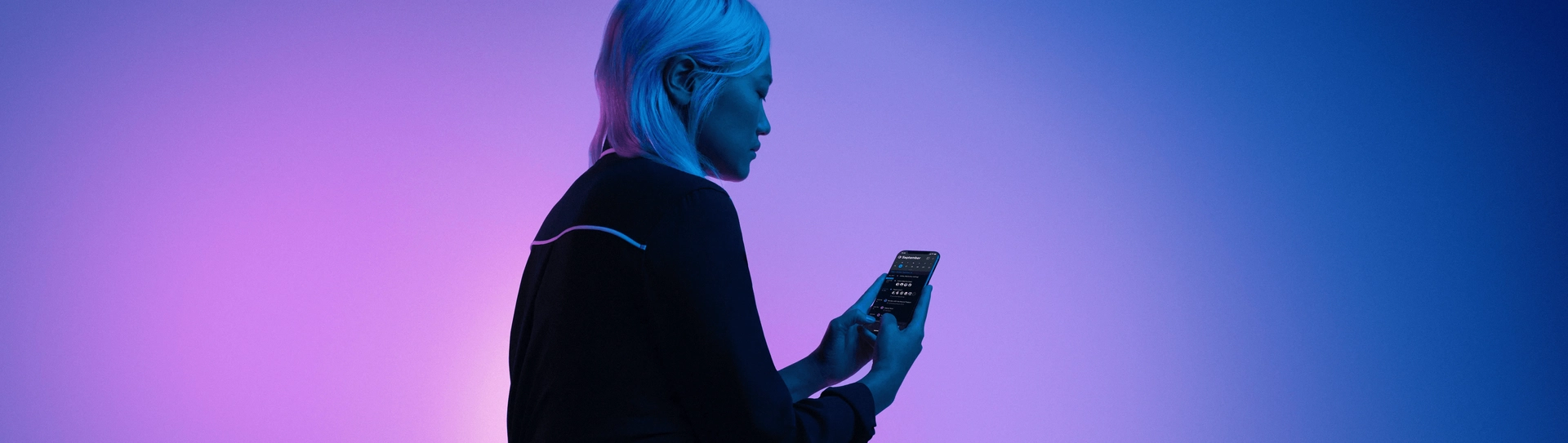 A person with short light blue hair stands against a gradient purple and blue background, looking at their smartphone, which emits a glow.