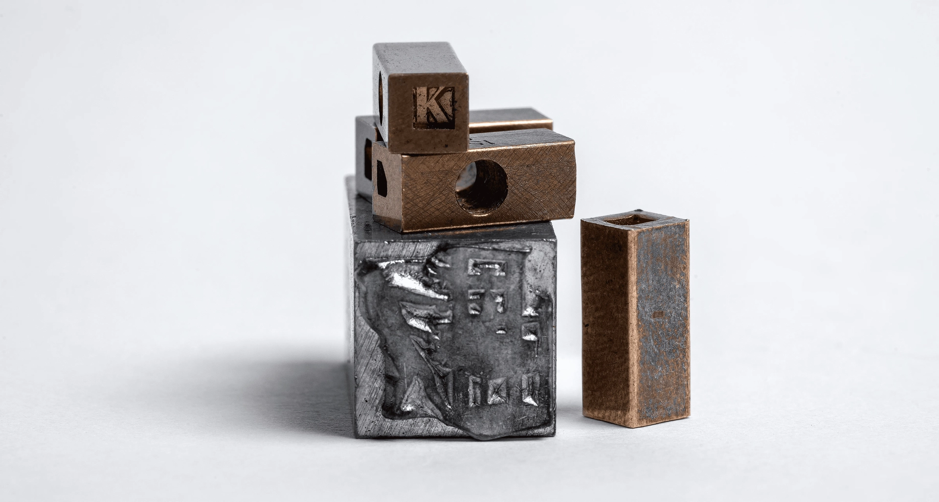 A close-up of several industrial metal blocks stacked on top of each other against a plain white background. One block has the letter 'K' engraved on it, while another block has a hole through it. The bottom block displays an engraved figure.