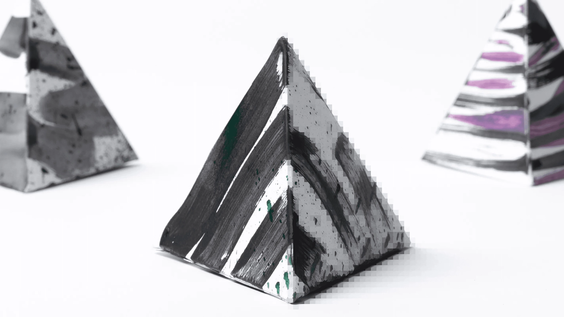 Three small abstract pyramids are displayed, each with distinct and artistic paint patterns. The foreground pyramid features a black and white striped design, while the background pyramids have a mix of marbled and striped colors, including purple accents.