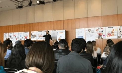 A speaker stands in front of a seated audience in a conference room with a series of posters displayed on the wall behind him. The audience members are attentively listening and taking notes. The posters contain various illustrations and text related to the presentation.