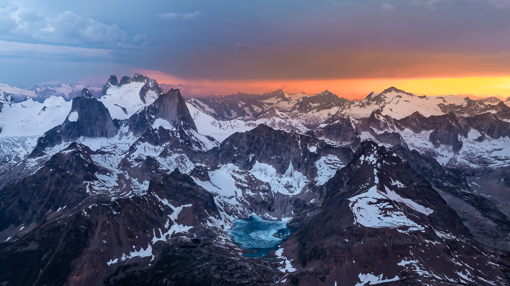 A dramatic sunset over a snow-capped mountain range with rugged peaks, perfect for wallpapers, features a small turquoise glacial lake in the foreground under a sky transitioning from blue to vibrant orange hues.