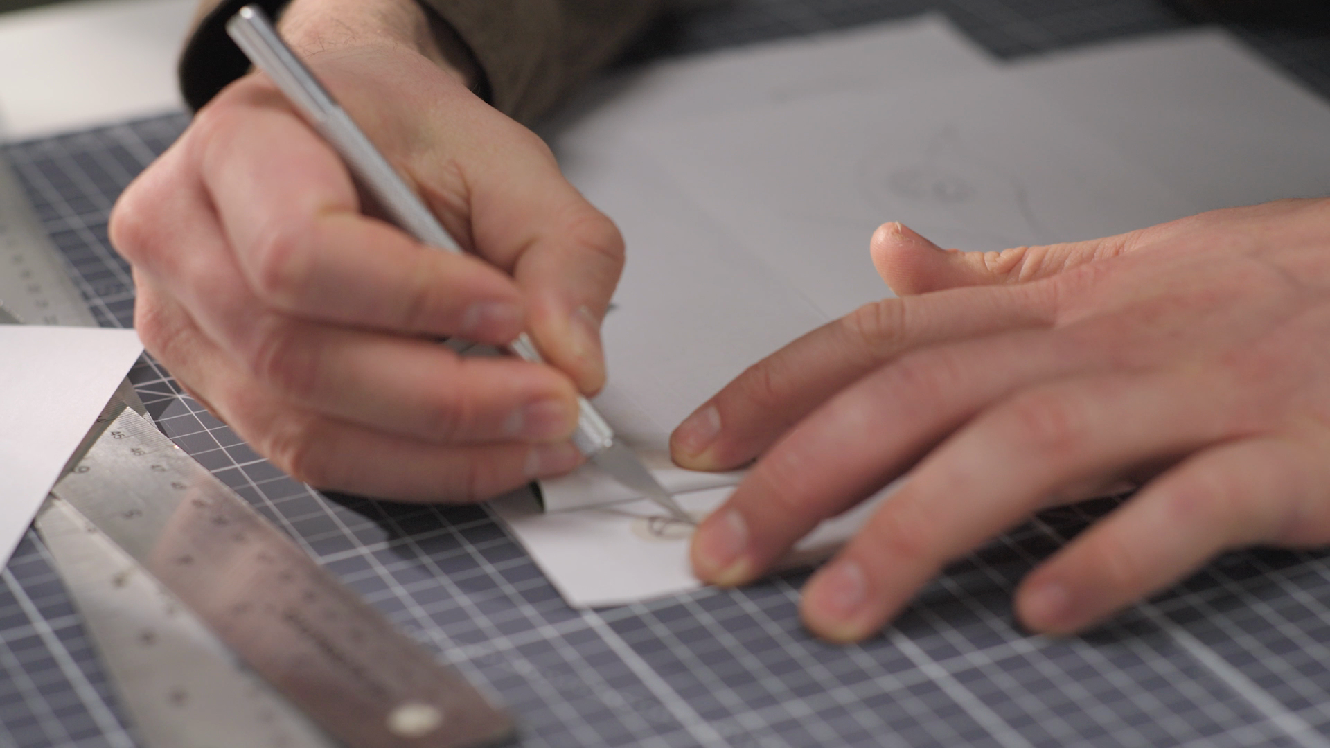 Close-up of hands using an X-acto knife to cut paper on a cutting mat. A metal ruler and another sheet of paper with a pencil sketch are visible.