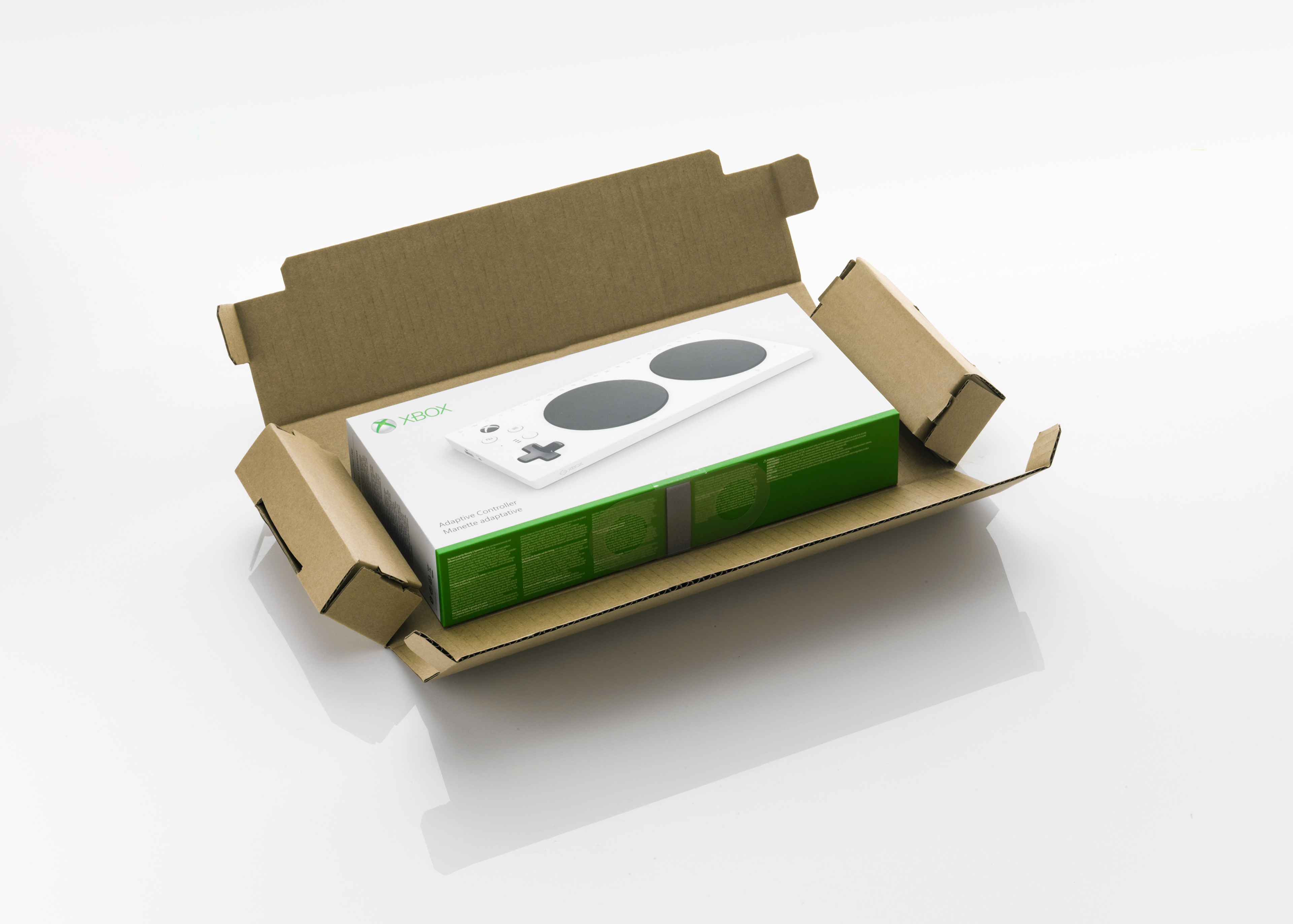 Xbox Adaptive Controller in its packaging is shown inside an open cardboard box. The controller’s white surface with two large black buttons is visible on the box's cover. The box rests on a white reflective surface.