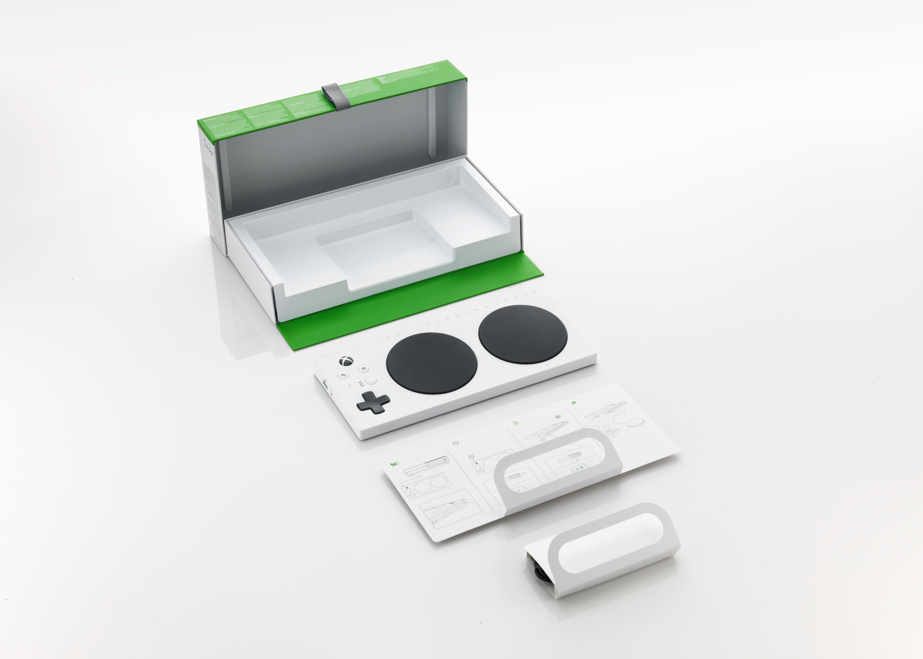 An Xbox Adaptive Controller unboxed and displayed. It includes a white rectangular controller with two large black circular buttons, a directional pad, and other buttons. A compact manual and packaging materials are also shown.