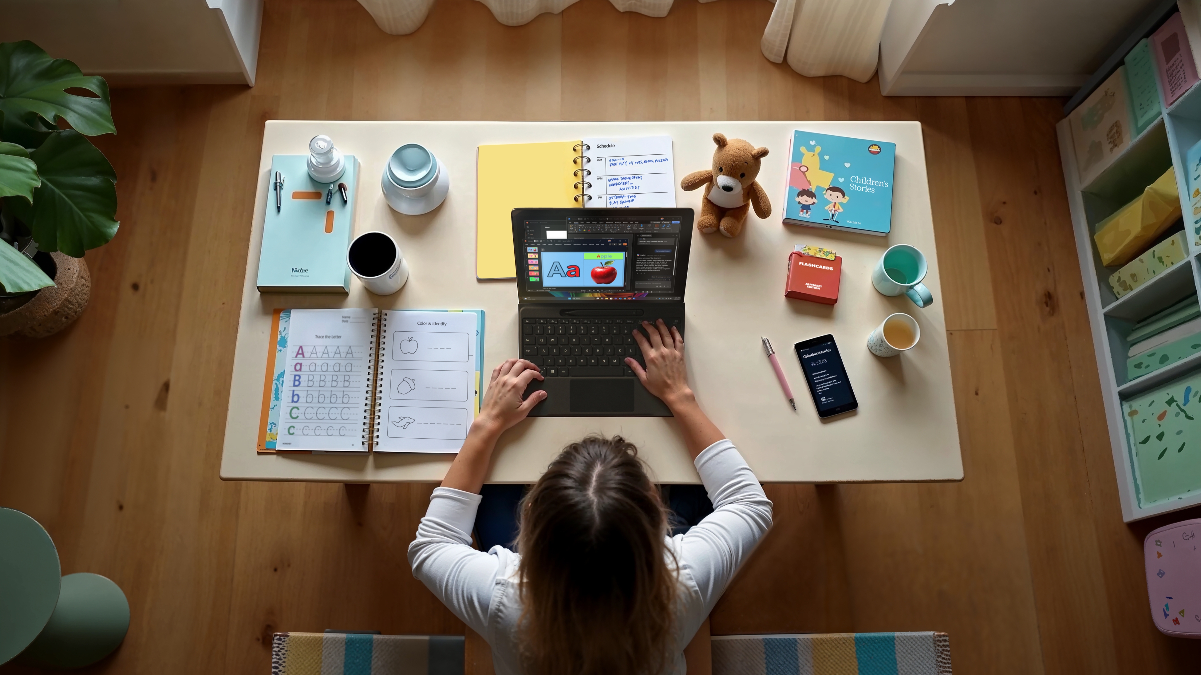 A woman sits at a desk working on a laptop surrounded by notebooks, a children's workbook, a smartphone, a teddy bear, and a cup, suggesting remote work or study in a home environment.
