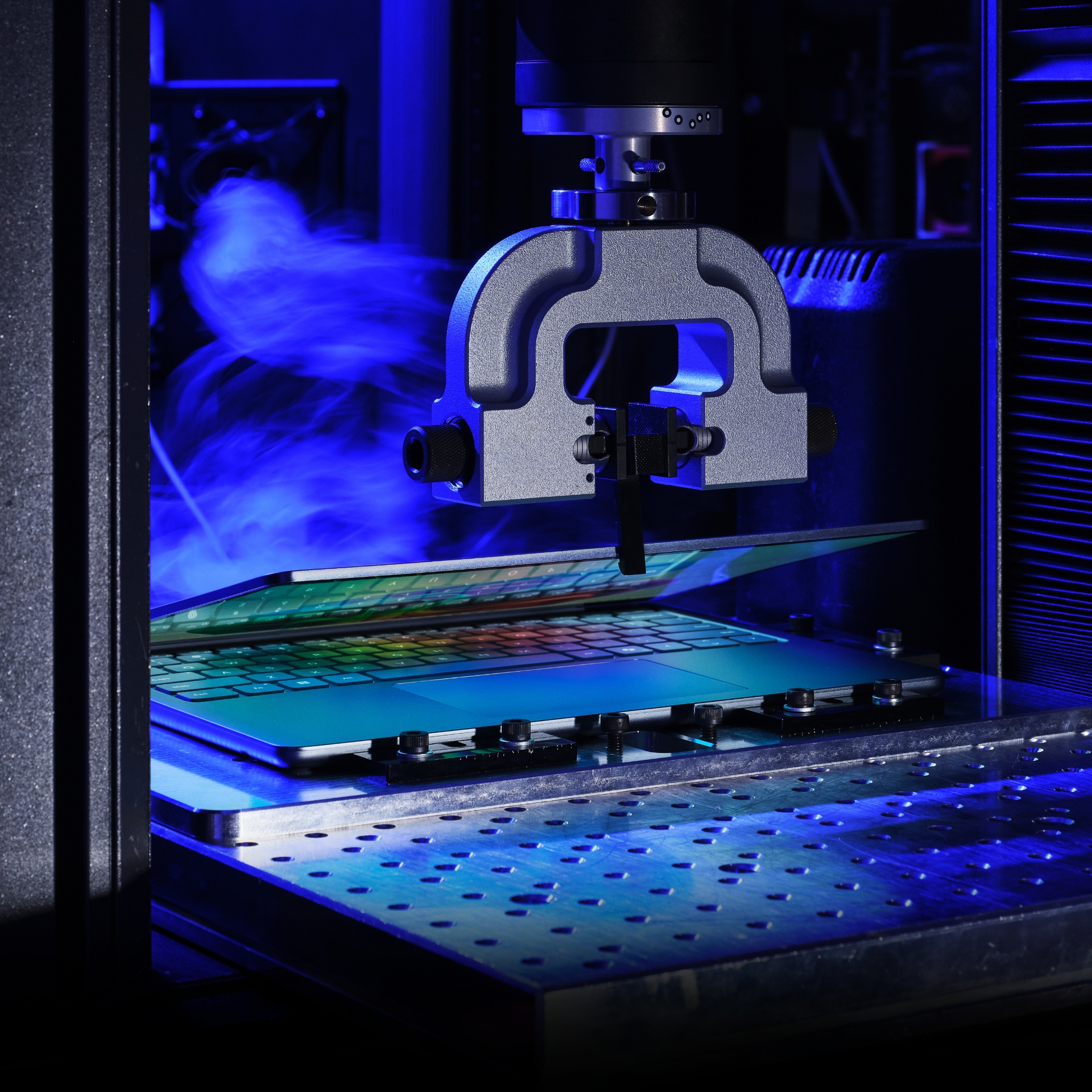 A precision machine arm tests or inspects a circuit board under blue lighting, with wisps of vapor or smoke in the background, in a high-tech laboratory setting.
