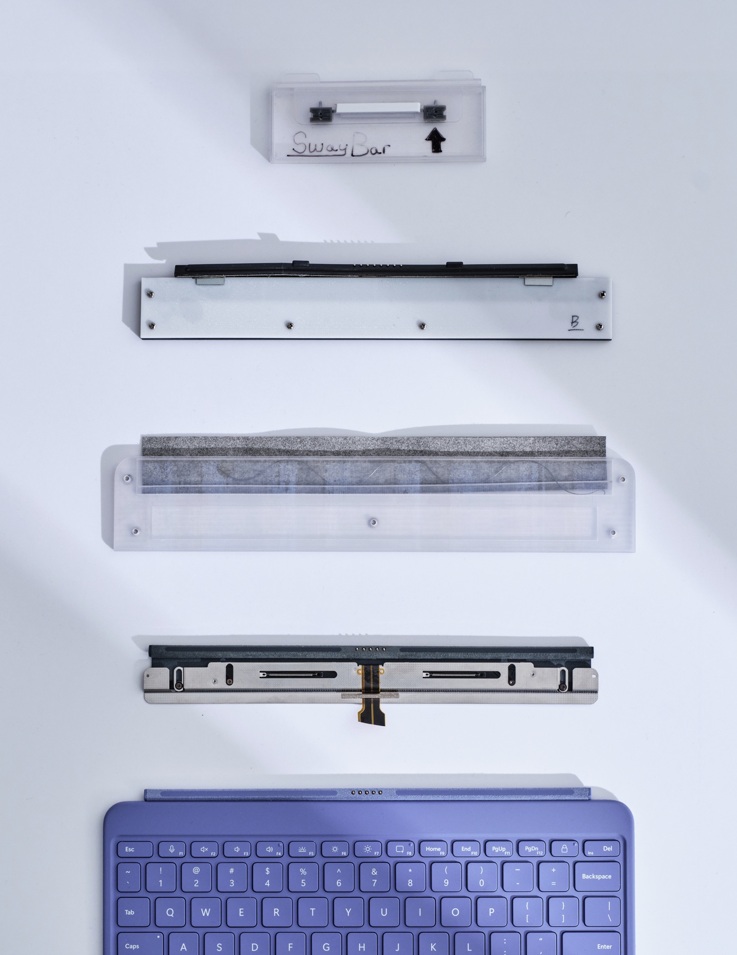Five computer hardware components, including a purple keyboard at the bottom, are arranged vertically on a white surface. The parts above the keyboard vary in size, color, and material, with some labeled and some containing metal pieces.