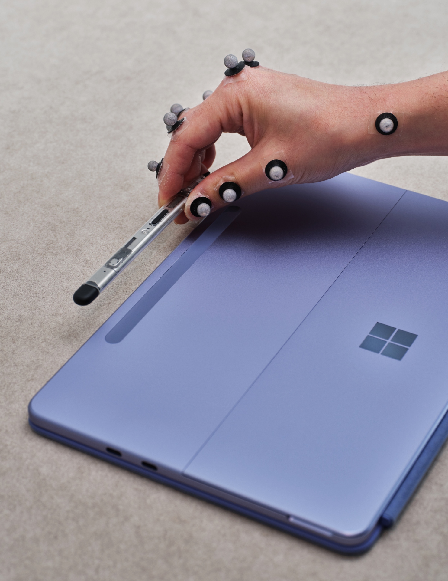 A hand with motion capture markers holds a stylus above a closed purple Microsoft Surface tablet on a beige surface.