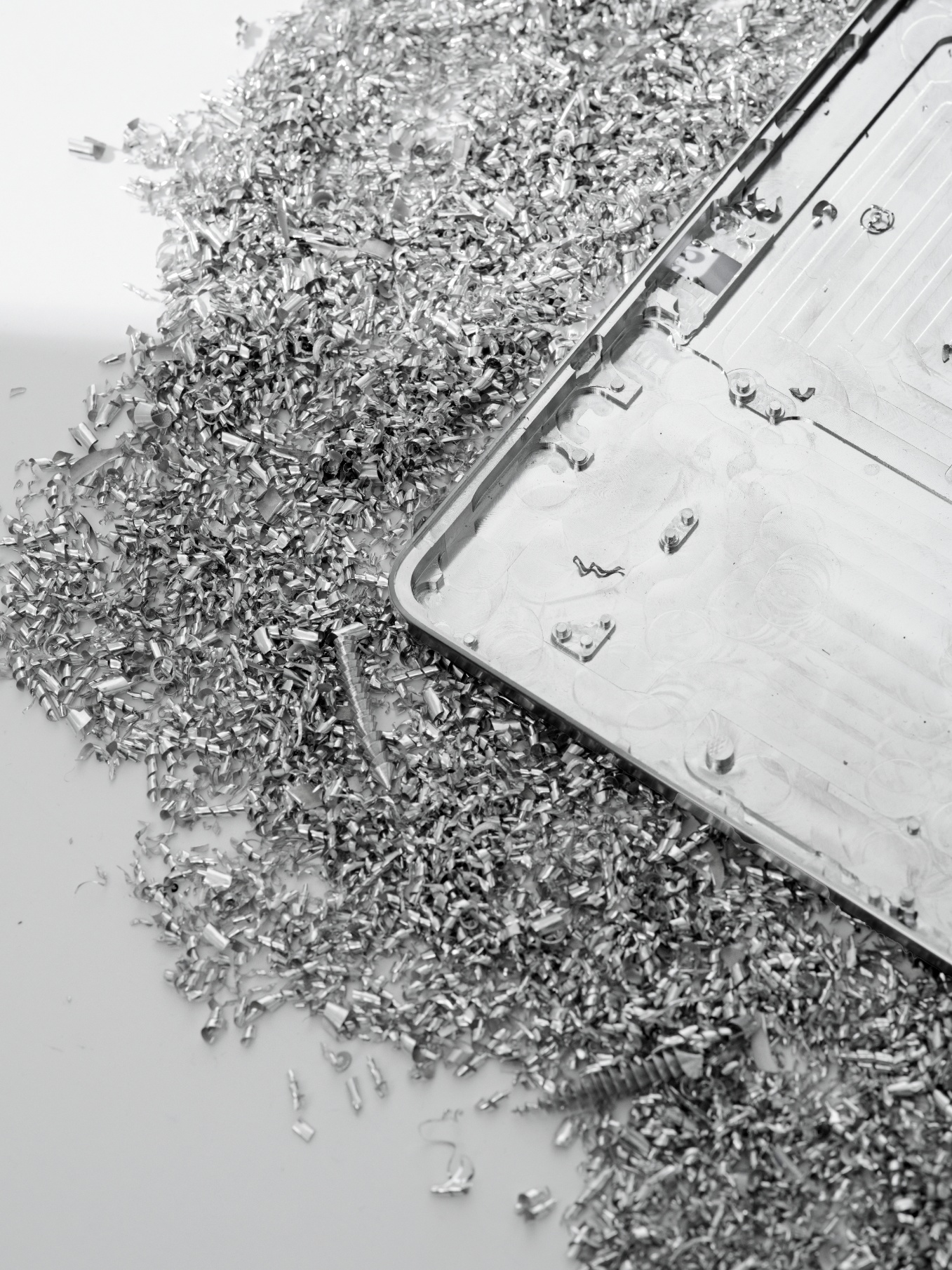 A metal sheet with grooves lies on a white surface, surrounded by scattered shiny metal shavings and fragments, suggesting recent machining or cutting.