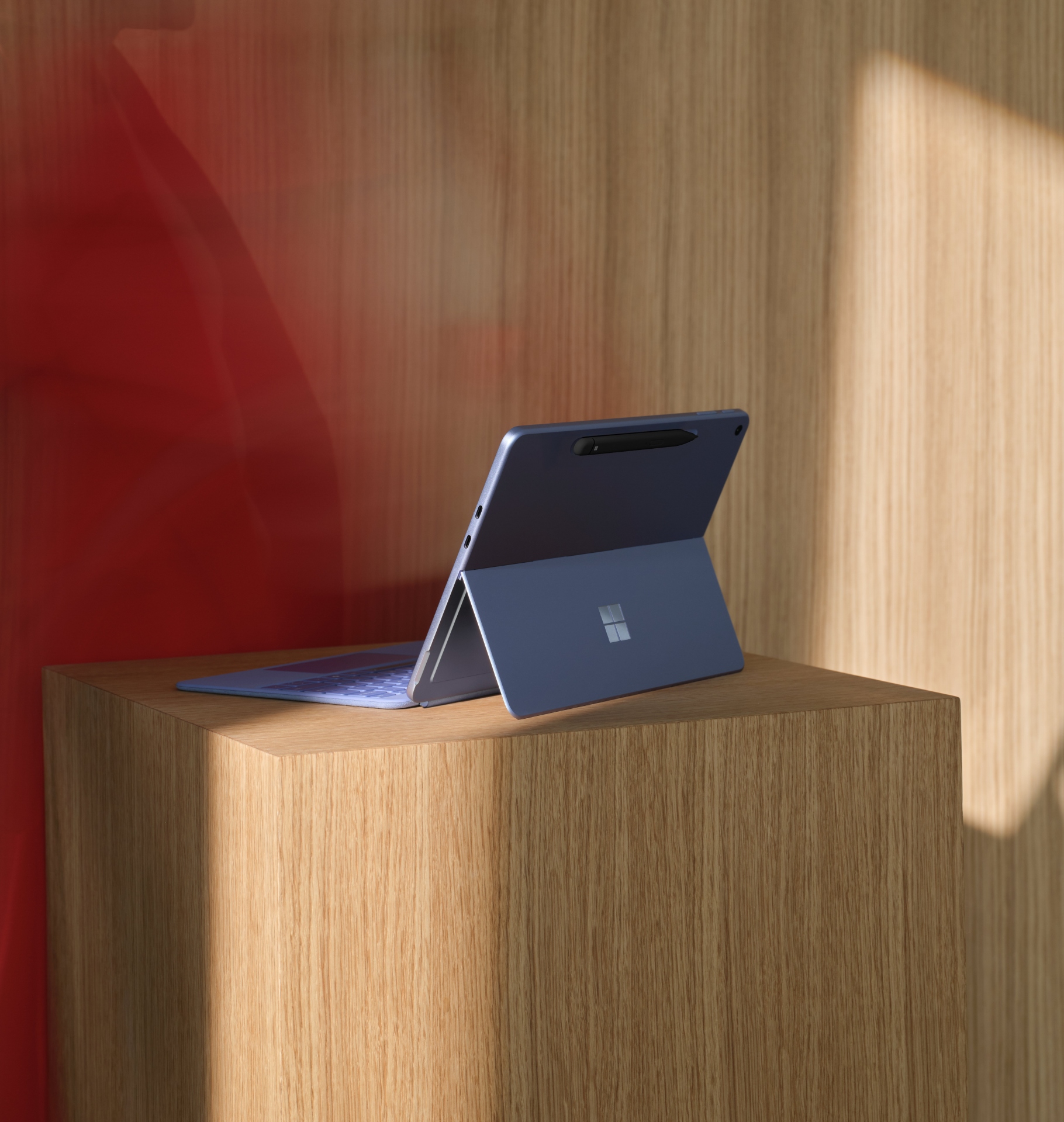 A Microsoft Surface tablet with a keyboard is propped up on a wooden pedestal, positioned against a wooden wall with a red reflection on the left side.