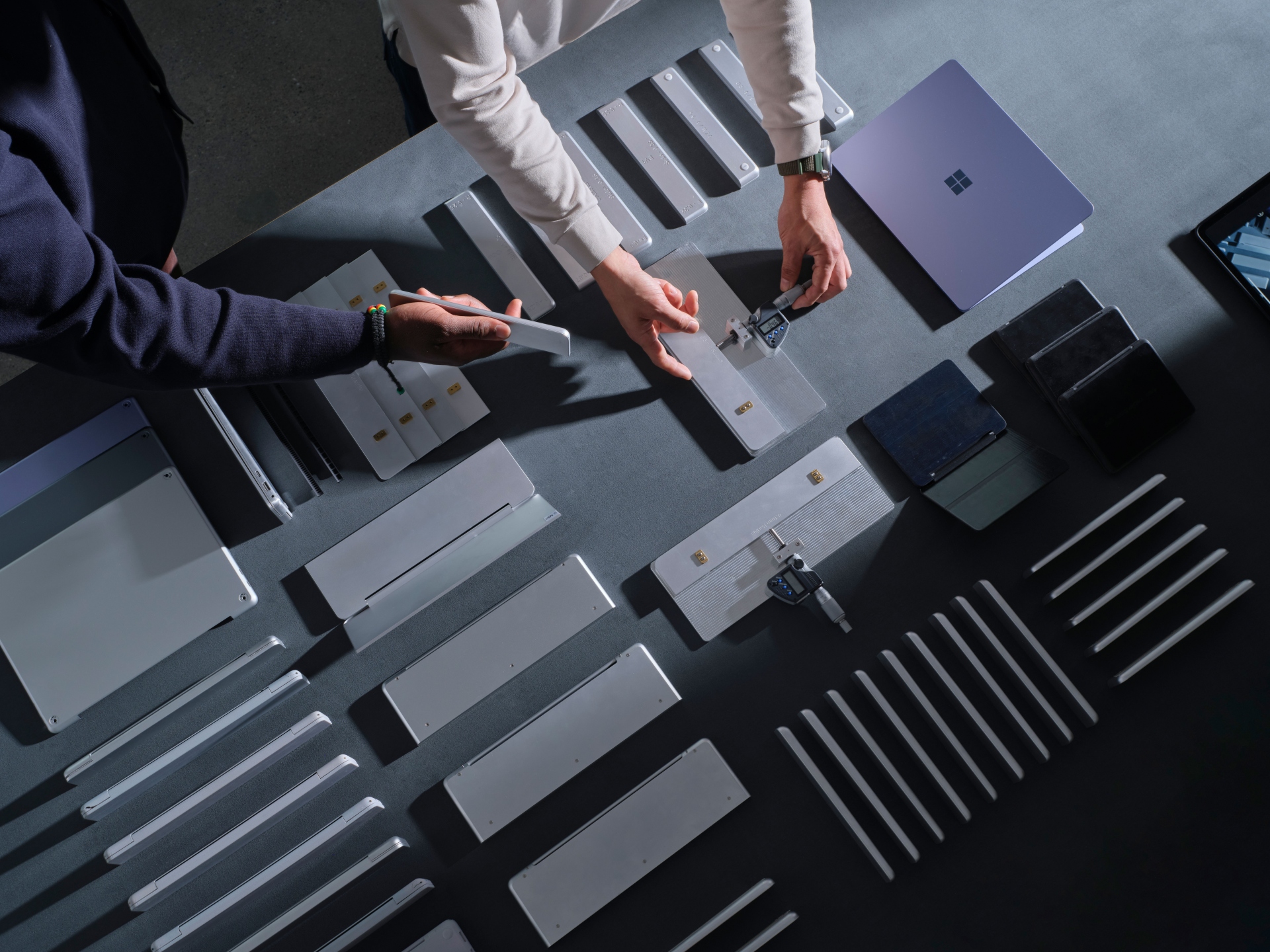 Two people arrange and examine metal and electronic components on a table, with a laptop nearby. The parts are organized in neat rows, suggesting a design or assembly process.