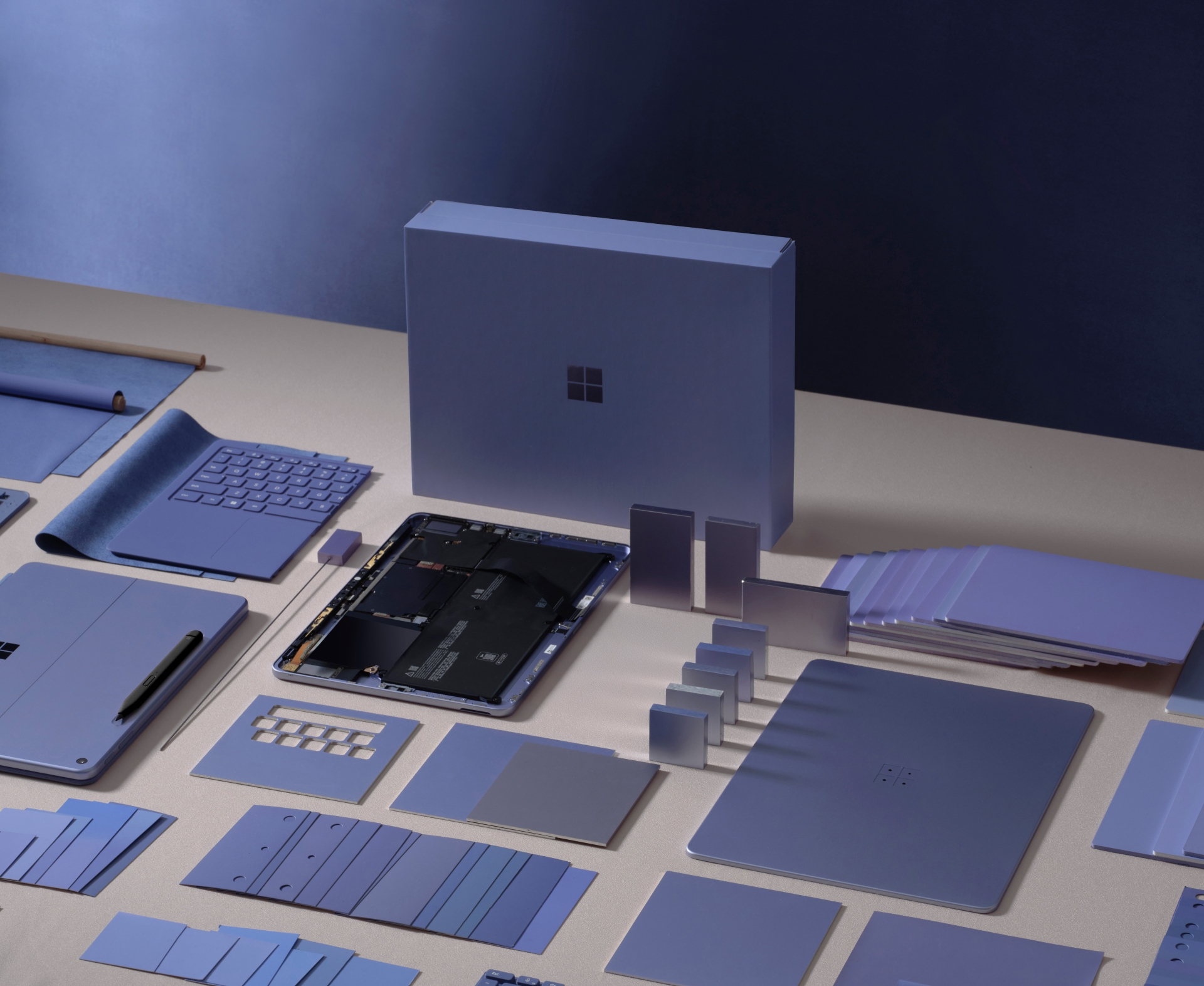 A collection of purple Microsoft Surface device parts and accessories, including keyboards, panels, circuit boards, and packaging, are neatly arranged on a beige surface against a blue background.