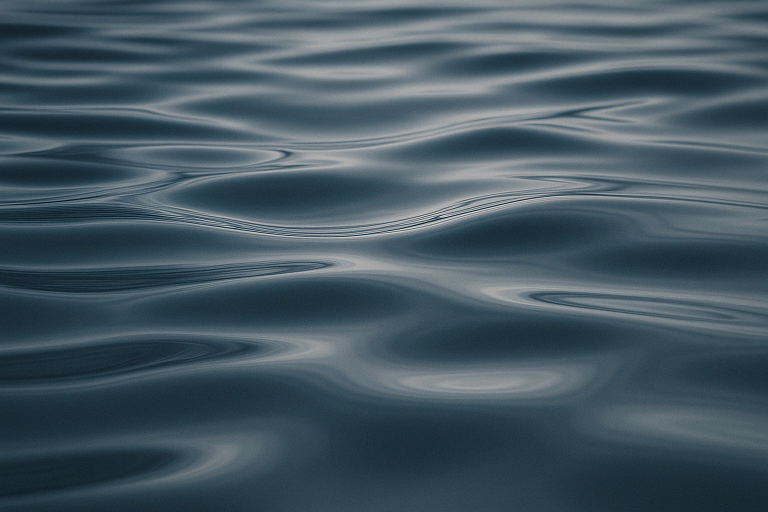 Close-up of gently rippling water, with soft reflections creating smooth, wavy patterns on the dark surface. The overall tone is cool and tranquil.