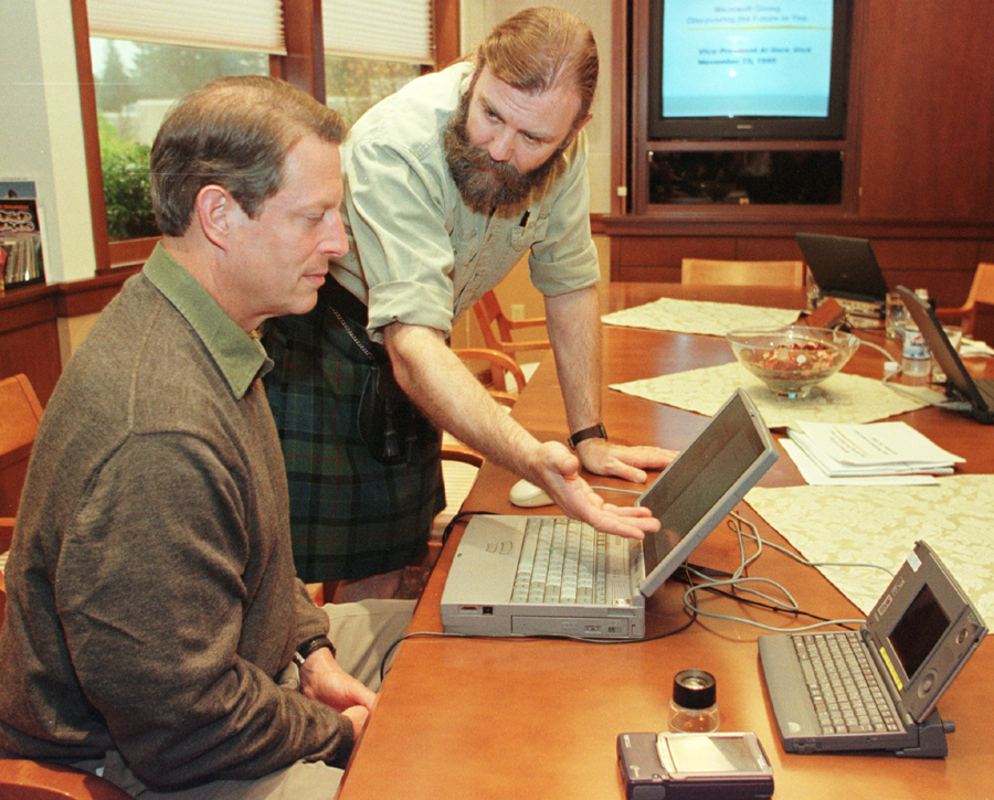 Two men in a conference room look at an open laptop on a wooden table. One is seated, and the other stands, gesturing toward the screen. Other laptops and electronic devices are also on the table.