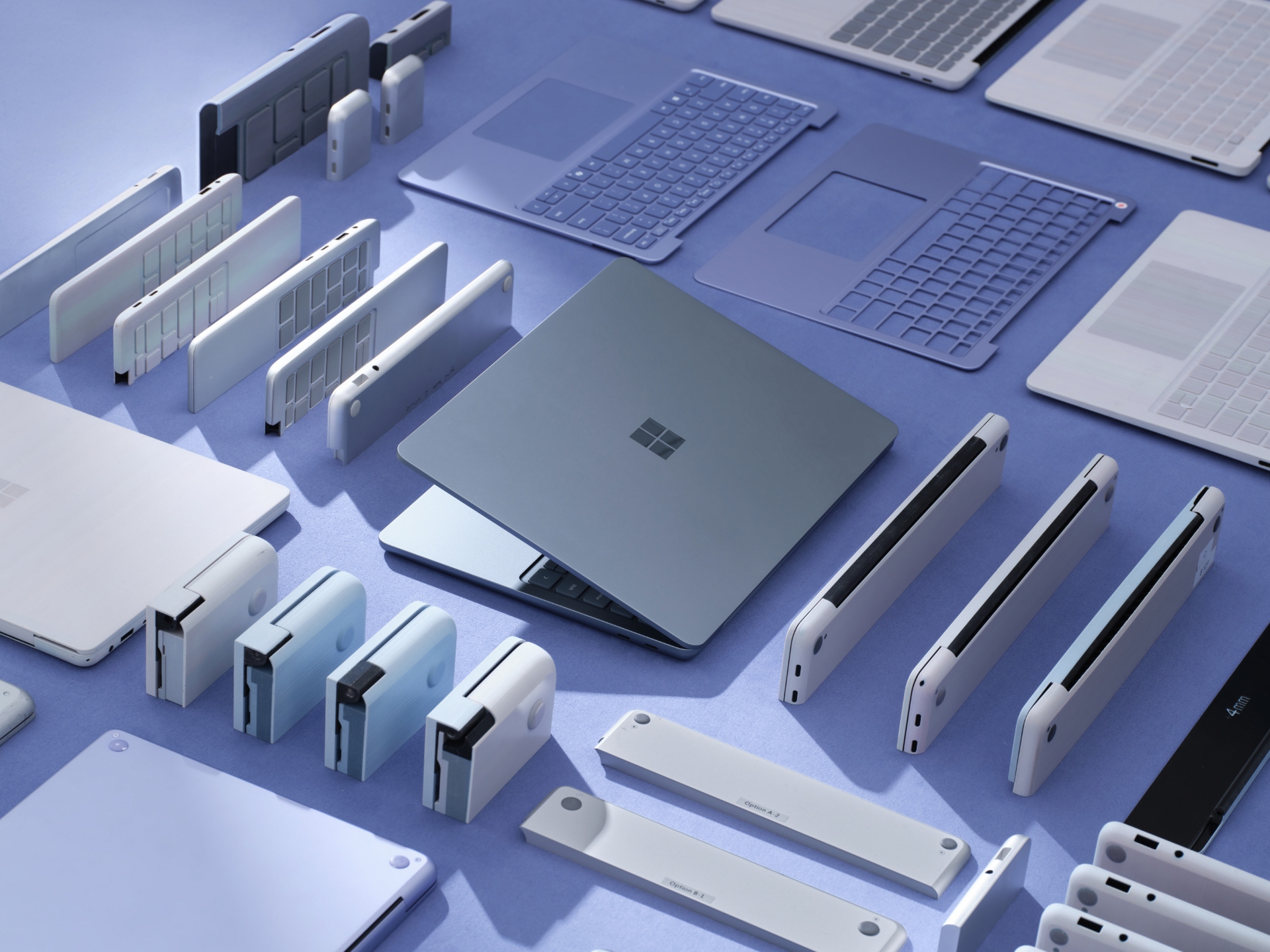 Various Microsoft Surface laptops, tablets, keyboards, and accessories are arranged neatly on a blue surface, showcasing different colors, components, and parts in a flat lay style.